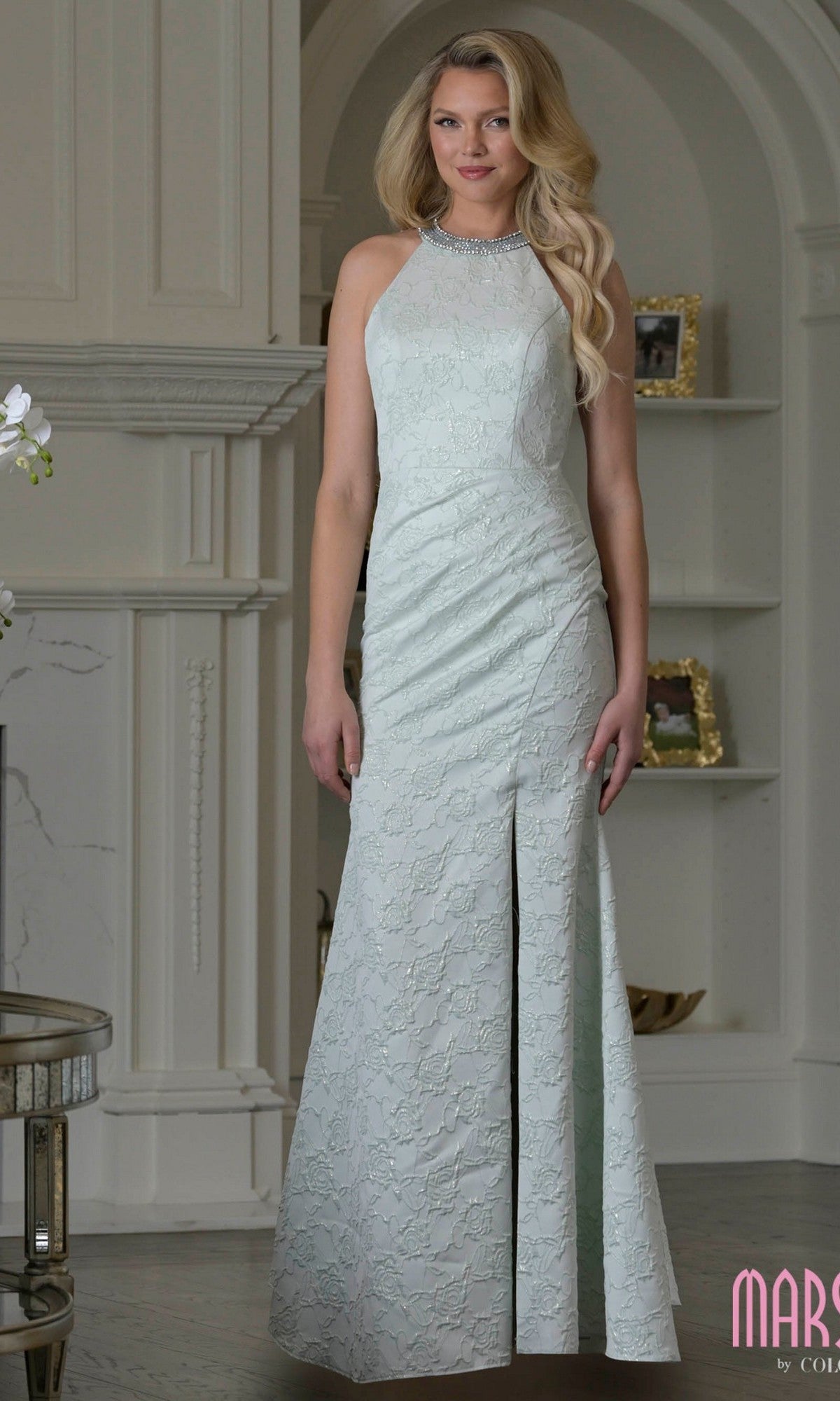 A woman stands indoors wearing the Marsoni M356—a long formal dress in light sage, featuring a sleeveless design, high neckline, lace detailing, and side slit.